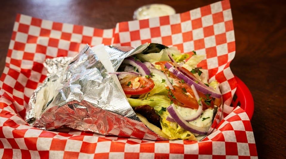 A gyro with lettuce, tomatoes, red onions, and sauce in a red and white checkered basket.