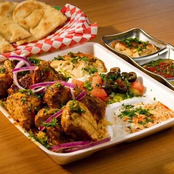 A takeout container with chicken, hummus, salad, and pita bread on a wooden table.