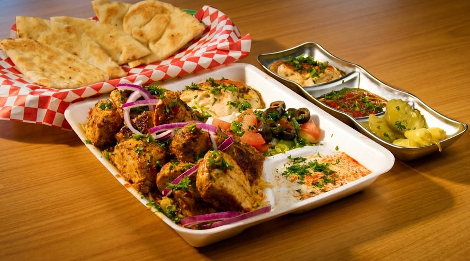 A takeout container with chicken, hummus, salad, and pita bread on a wooden table.