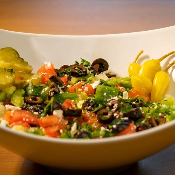 A white bowl filled with a colorful salad of chopped vegetables, olives, and pickles.