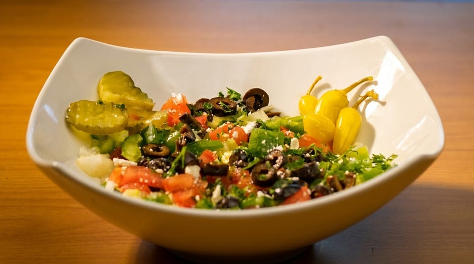 A white bowl filled with a colorful salad featuring olives, tomatoes, peppers, and pickles.
